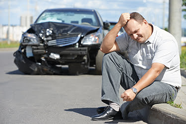 Man sitting on the curb in front of car with a crumpled front end after an accident