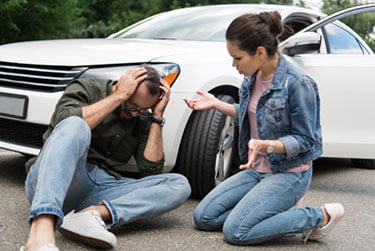 Man sitting in front of a car with his head in his hands and woman kneeling next to him after a pedestrian accident