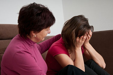 Older woman in a rose colored long sleeve shirt comforting a younger woman in a red short sleeve shirt with her head in her hands as they discuss a wrongful death case